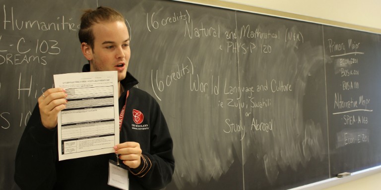 Peer coach leading a group workshop showing a worksheet and standing in front of a blackboard with notes about scheduling.